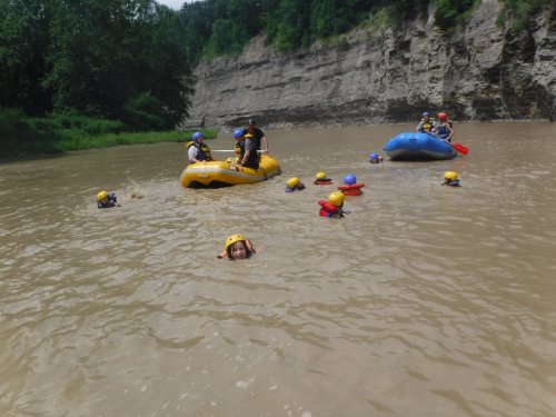 Group of people in helmets rafting and swimming in a river, with rocky cliffs and greenery in the background.