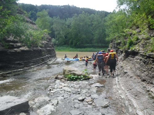 A group of people carrying rafts along a rocky riverbank surrounded by lush greenery and hills.