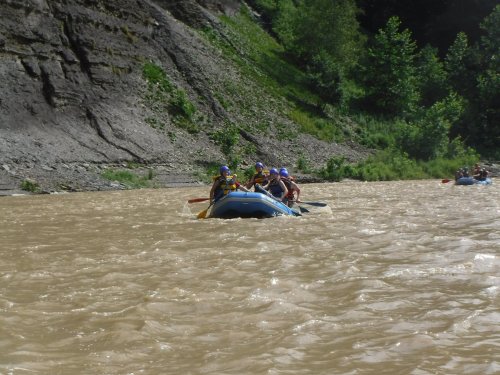 A group of people in a blue raft navigating a muddy river surrounded by steep, green banks.