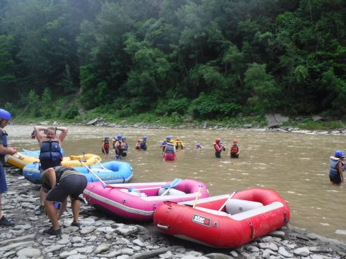 Group of people in life jackets near rafts on a riverbank, some wading in the water, surrounded by lush greenery.
