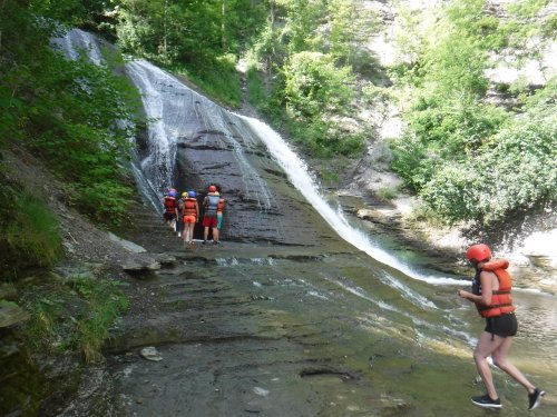 A group of people in life jackets approaches a waterfall in a lush, green forested area. One person walks nearby.