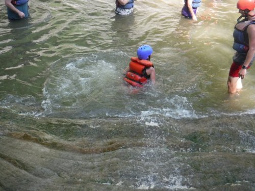 A child in an orange life jacket splashes in shallow water, surrounded by people wearing life vests.