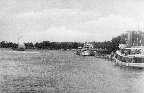 Black and white image of a river with boats docked and a sailboat in the distance, surrounded by trees and clouds.