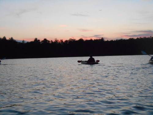A silhouette of a person kayaking on a calm lake at sunset, with trees lining the shore in the background.