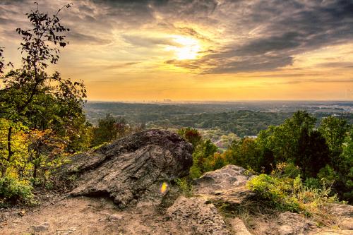 A scenic view from a rocky overlook, showcasing a sunset over a lush valley and distant city skyline.