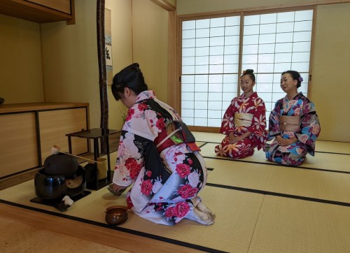 Three women in colorful kimonos participate in a traditional tea ceremony in a serene, tatami-matted room.