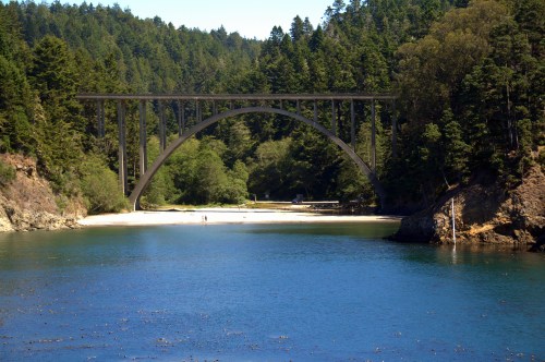 A scenic view of a large arch bridge over a calm river, surrounded by lush green trees and a sandy beach.