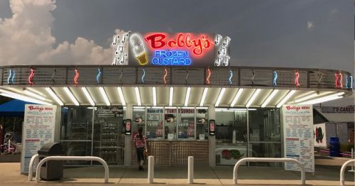 A colorful ice cream shop with neon signs, featuring a menu and outdoor seating under a cloudy sky.