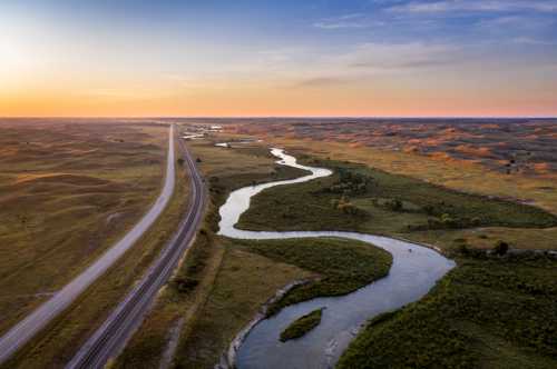 Aerial view of a winding river alongside a highway, surrounded by rolling hills and a colorful sunset sky.