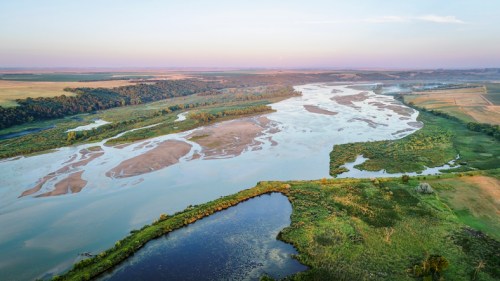 Aerial view of a winding river surrounded by lush greenery and fields under a soft, pastel sky.