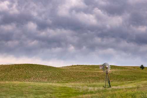 A windmill stands on a grassy hill under a cloudy sky, with rolling green fields in the background.