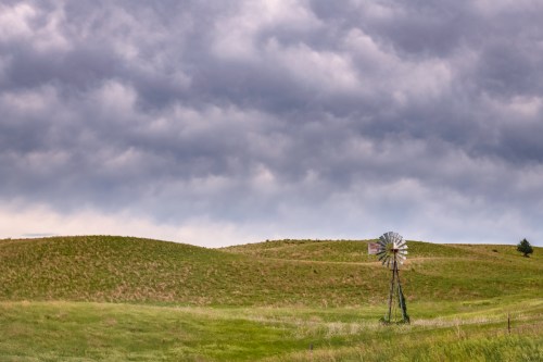 A windmill stands on a grassy hill under a cloudy sky, with rolling green fields in the background.
