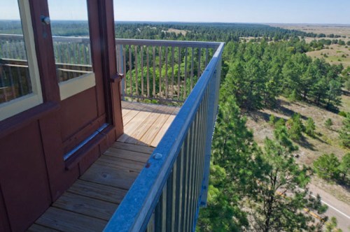 View from a lookout tower balcony overlooking a forested landscape and open fields under a clear blue sky.