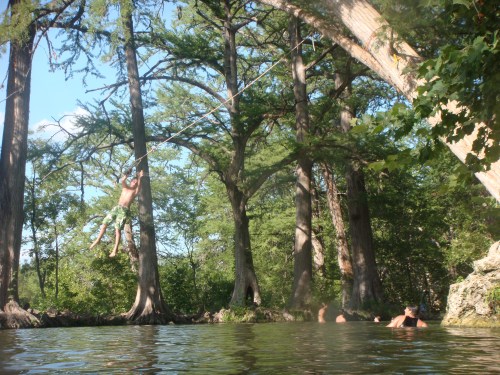 A person swings from a rope into a natural swimming hole surrounded by tall trees and others enjoying the water.