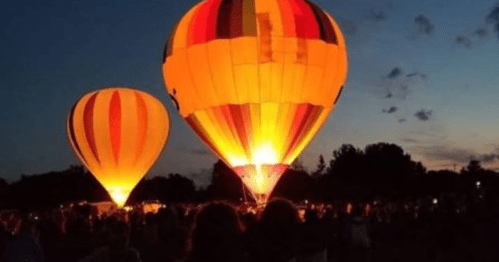 Two brightly colored hot air balloons glow against a twilight sky, surrounded by a crowd of spectators.