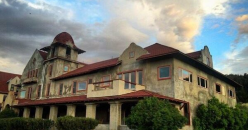 A large, weathered mansion with a red roof, surrounded by greenery and under a cloudy sky.
