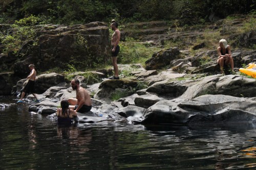 A group of people relaxes by a rocky riverbank, some sitting on rocks and others wading in the water.