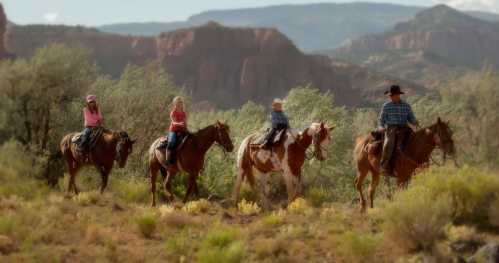 Four people on horseback ride through a scenic landscape with mountains and greenery in the background.