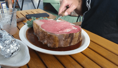 A person slices into a large, juicy prime rib on a white plate, with sides and a phone nearby on a wooden table.