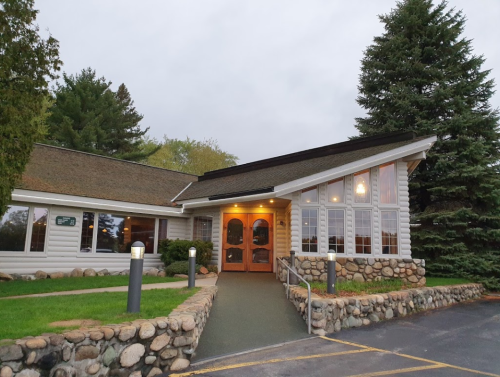 A cozy, modern building with large windows, stone accents, and a welcoming entrance surrounded by greenery.