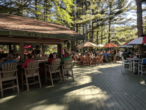 A lively outdoor dining area with people seated at tables under umbrellas, surrounded by trees.