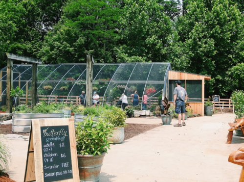 A butterfly greenhouse with visitors, surrounded by greenery and potted plants, featuring a sign with admission prices.