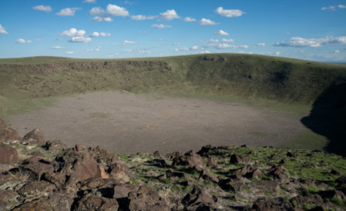 A vast, empty crater surrounded by rocky terrain and green hills under a blue sky with scattered clouds.