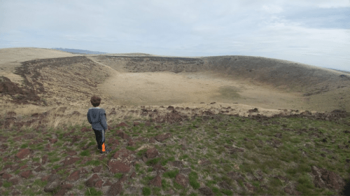 A child stands on rocky terrain, gazing at a large, grassy crater under a cloudy sky.