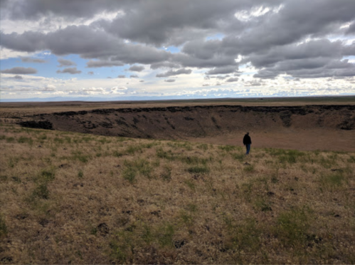 A person walks along the edge of a large crater, surrounded by grassy terrain and a cloudy sky.
