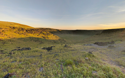 A vast, grassy landscape with a large, circular crater under a clear sky at sunset.