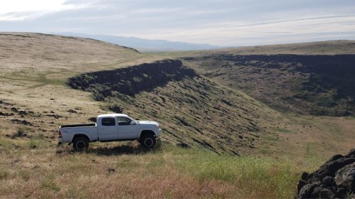 A white pickup truck parked on a grassy hillside overlooking a steep, rocky cliff and distant landscape.