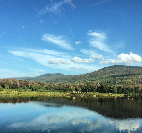 A serene lake surrounded by green hills and colorful trees under a blue sky with fluffy clouds.