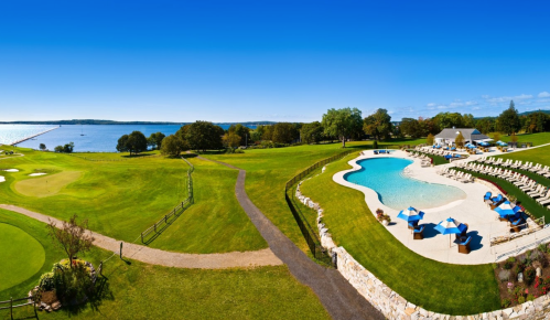 A scenic view of a golf course and pool area by a lake, with blue skies and lounge chairs around the pool.