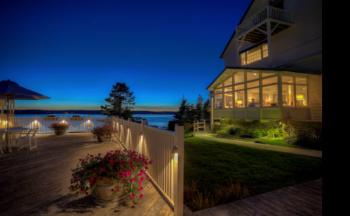 A serene evening view of a waterfront house with outdoor seating and vibrant flowers, illuminated by soft lights.