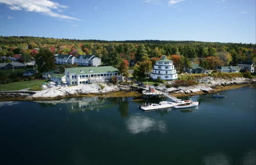 A scenic waterfront view featuring a white building, boats, and colorful trees along the shore under a clear blue sky.