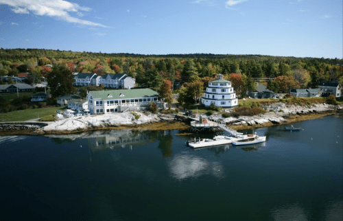 A scenic waterfront view featuring a white building, boats, and colorful trees along the shore under a clear blue sky.