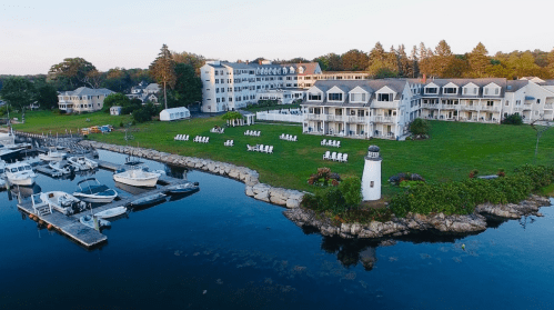 A scenic waterfront view of a resort with boats docked, a lighthouse, and green lawns under a clear sky.