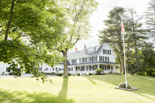 A large, white house with a dark roof, surrounded by trees and a grassy lawn, featuring an American flag on a pole.