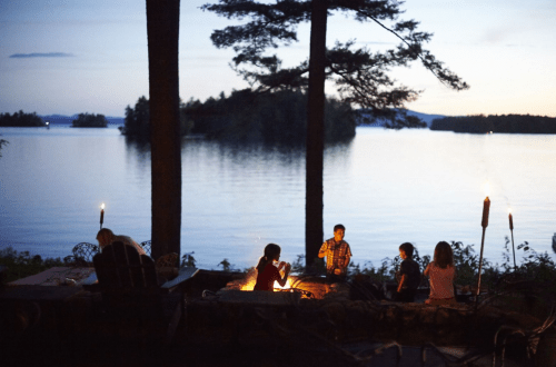 A serene lakeside scene at dusk with children gathered around a campfire, surrounded by trees and water.