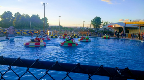 Colorful bumper boats float in a pool at sunset, with people enjoying the ride and a fence in the foreground.