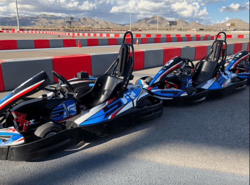 Three go-karts lined up on a racetrack with red and gray barriers and mountains in the background under a blue sky.