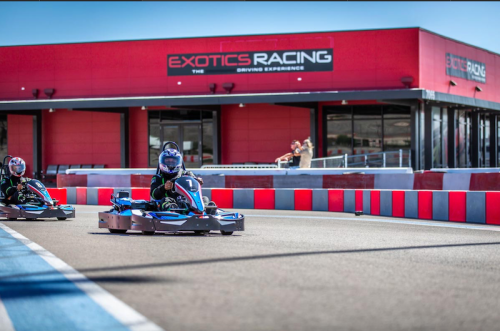 Two go-karts race on a track in front of a red building with "Exotics Racing" signage. Spectators watch in the background.