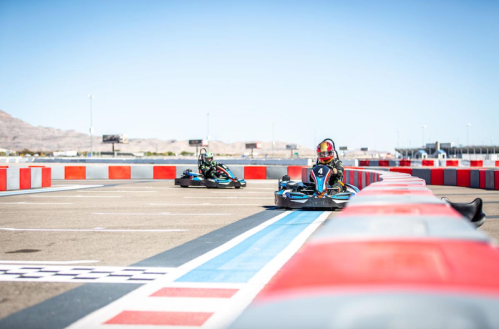 Two go-karts racing on a track with red and white barriers under a clear blue sky.