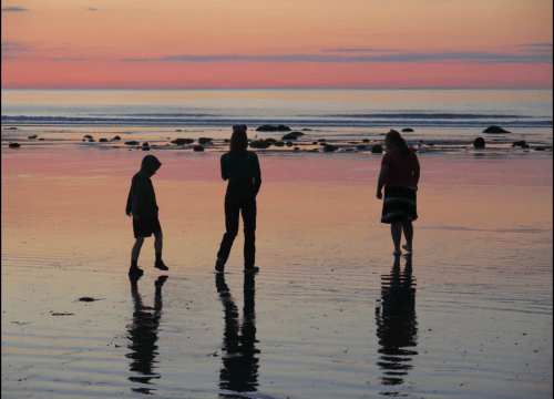 Silhouettes of three people walking on a beach at sunset, with colorful skies and reflections on the wet sand.
