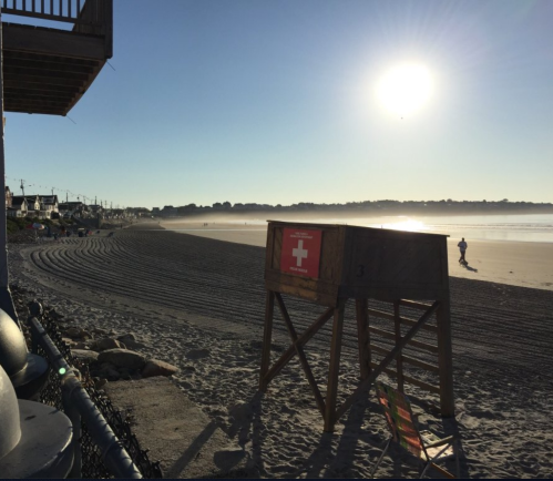 A sunny beach scene with a lifeguard stand, smooth sand, and a distant figure walking along the shore.
