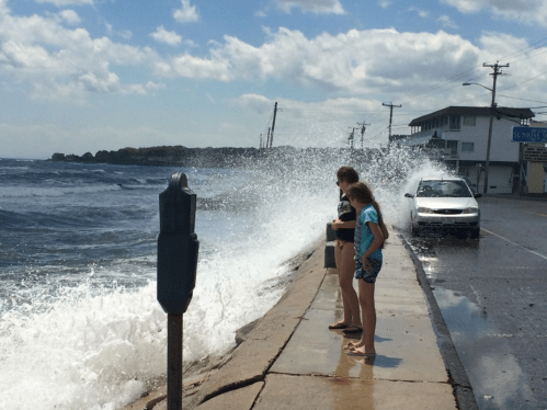 Two children stand on a waterfront sidewalk, watching waves crash against the shore, with a car parked nearby.