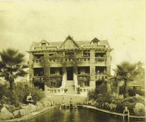 Historic building surrounded by lush greenery and a pond, with people relaxing by the water in a vintage setting.