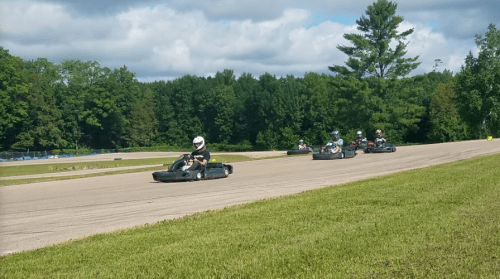 A group of go-karts racing on a track surrounded by green trees under a partly cloudy sky.