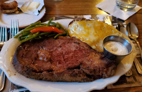 A plate featuring a juicy prime rib, green beans, cheesy potato, and a small cup of sauce, set on a wooden table.