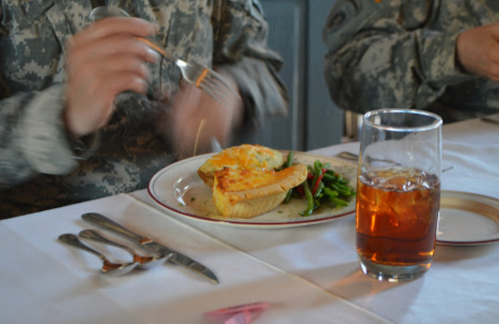 A soldier's hand holds a fork above a plate with a slice of quiche and green beans, next to a glass of iced tea.